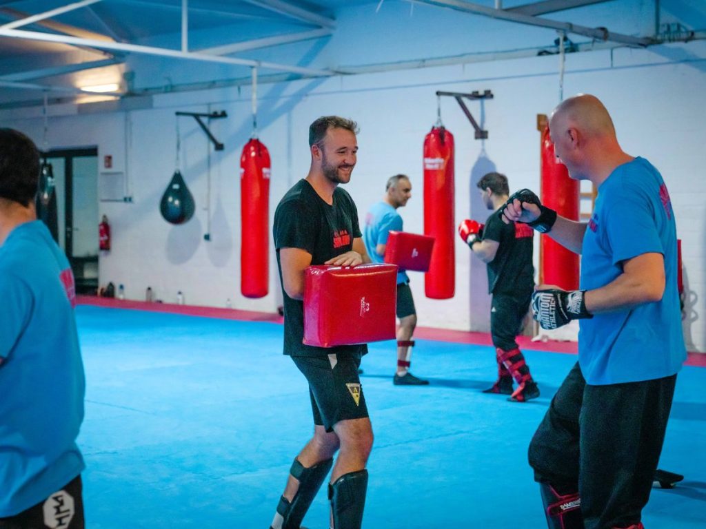 Gürtel und Graduierungen im Kampfsport: Was sie bedeuten 2 Lächelnder Schüler trägt ein rotes Kickpad, im Hintergrund trainieren weitere Mitglieder an den Boxsäcken.