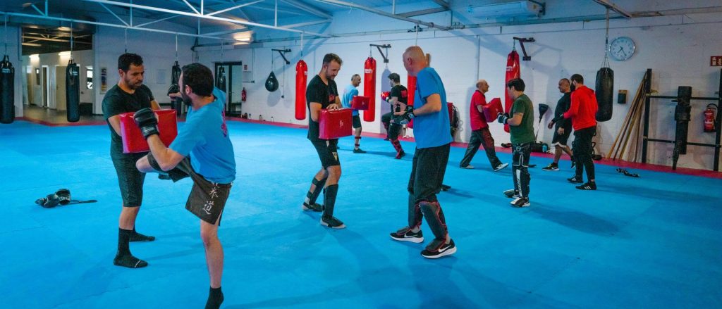 Weitwinkelaufnahme der Trainingshalle: acht Personen trainieren paarweise mit Pratzen auf der blauen Matte.
