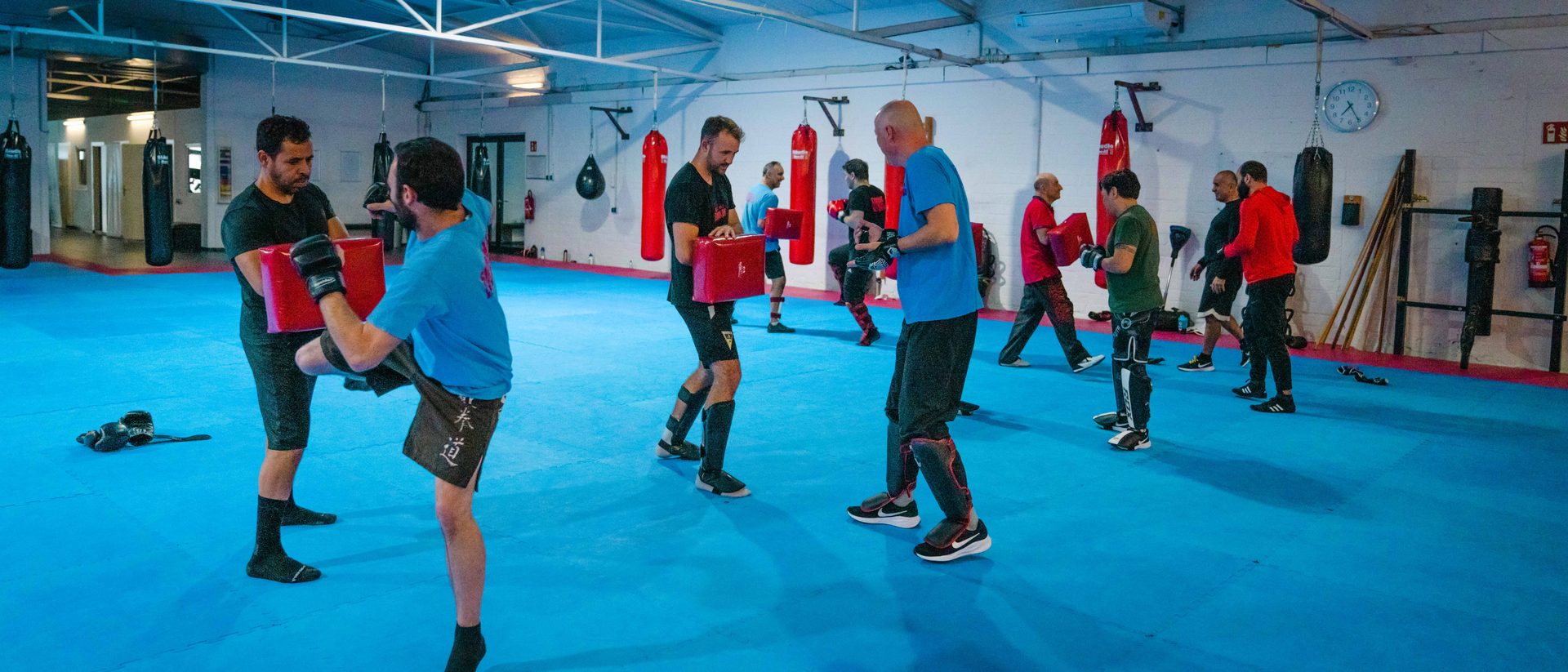 Weitwinkelaufnahme der Trainingshalle: acht Personen trainieren paarweise mit Pratzen auf der blauen Matte.