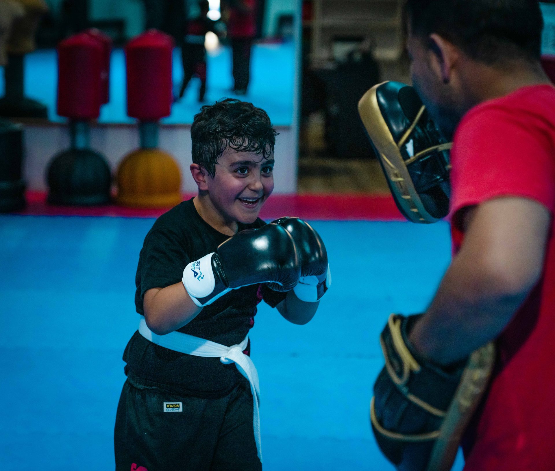Lachender Junge mit Boxhandschuhen trifft die Pratze seines Trainers im Kindertraining.