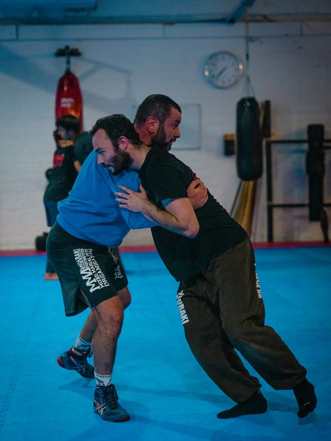 Zwei Trainierende im Ringen-Clinch vor Boxsack und Wanduhr der Trainingshalle.