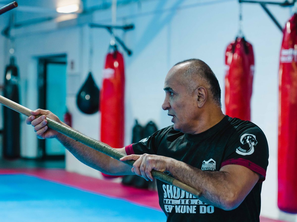 Der Sifu in dynamischer Haltung mit langem Holzstock vor einer Reihe roter Boxsäcke.