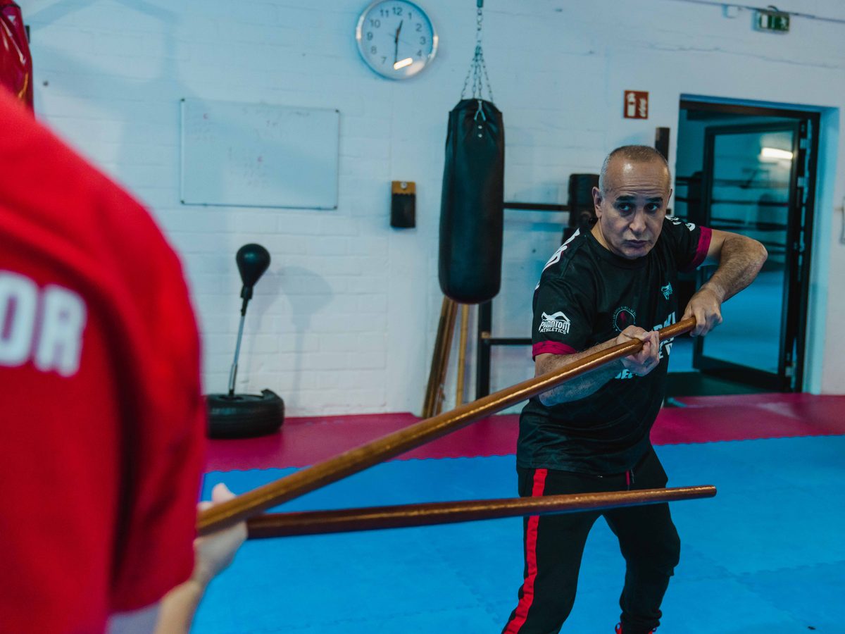 Der Sifu greift mit dem langen Stock den Instructor im roten Shirt an, Weitwinkelblick in die Halle.