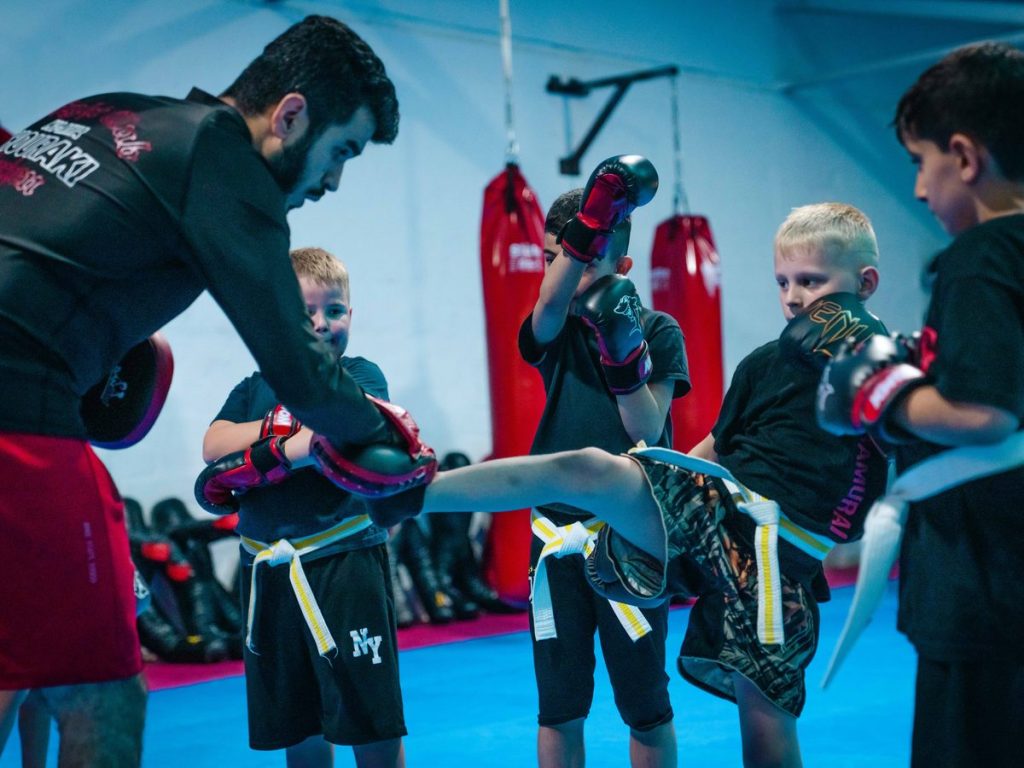 Kindergruppe im Halbkreis: Der Trainer hilft einem Kind bei einem hohen Kick, die anderen schauen zu.