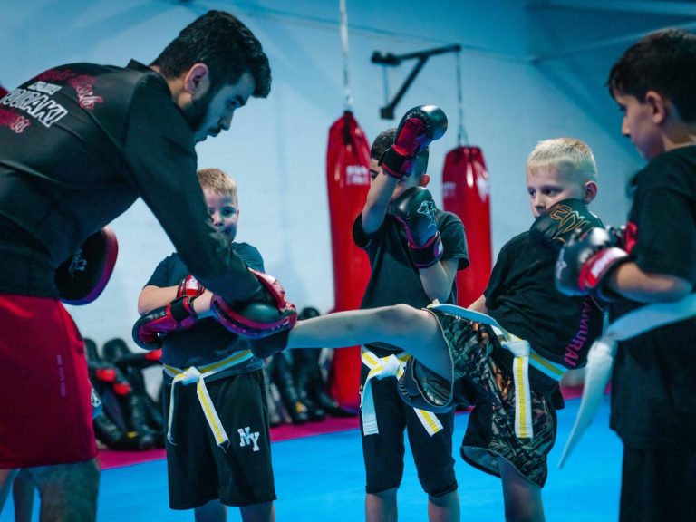 Kindergruppe im Halbkreis: Der Trainer hilft einem Kind bei einem hohen Kick, die anderen schauen zu.