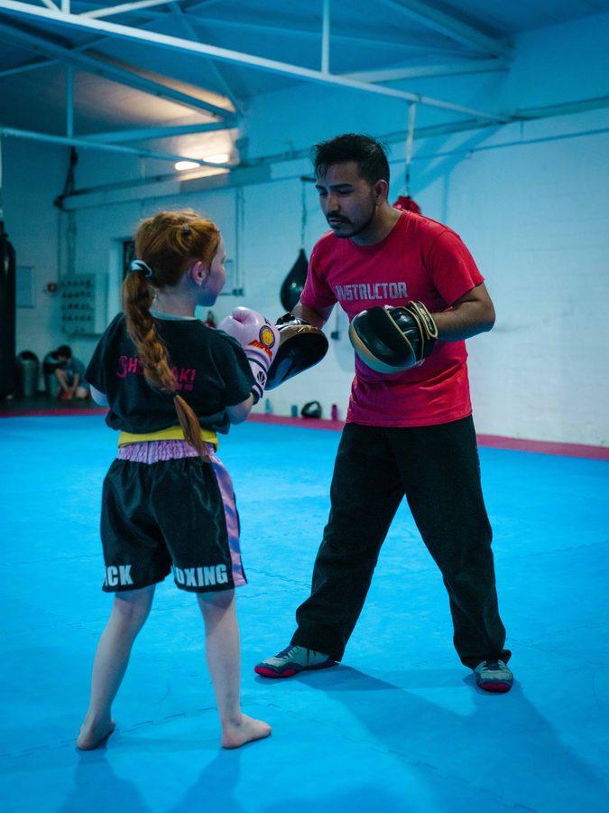 Mädchen mit rotem Zopf im schwarzen Kickbox-Outfit schlägt die Pratze des Trainers.
