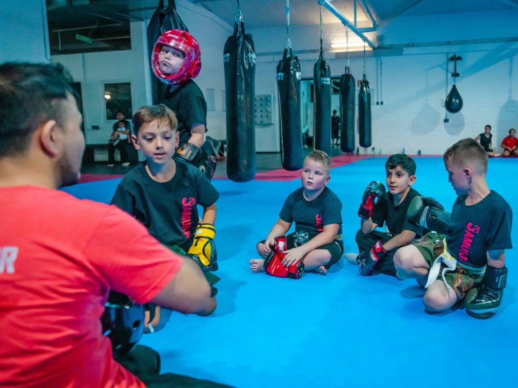 Kinder sitzen im Kreis auf der Matte, während der Trainer im roten Shirt mit ihnen spricht.