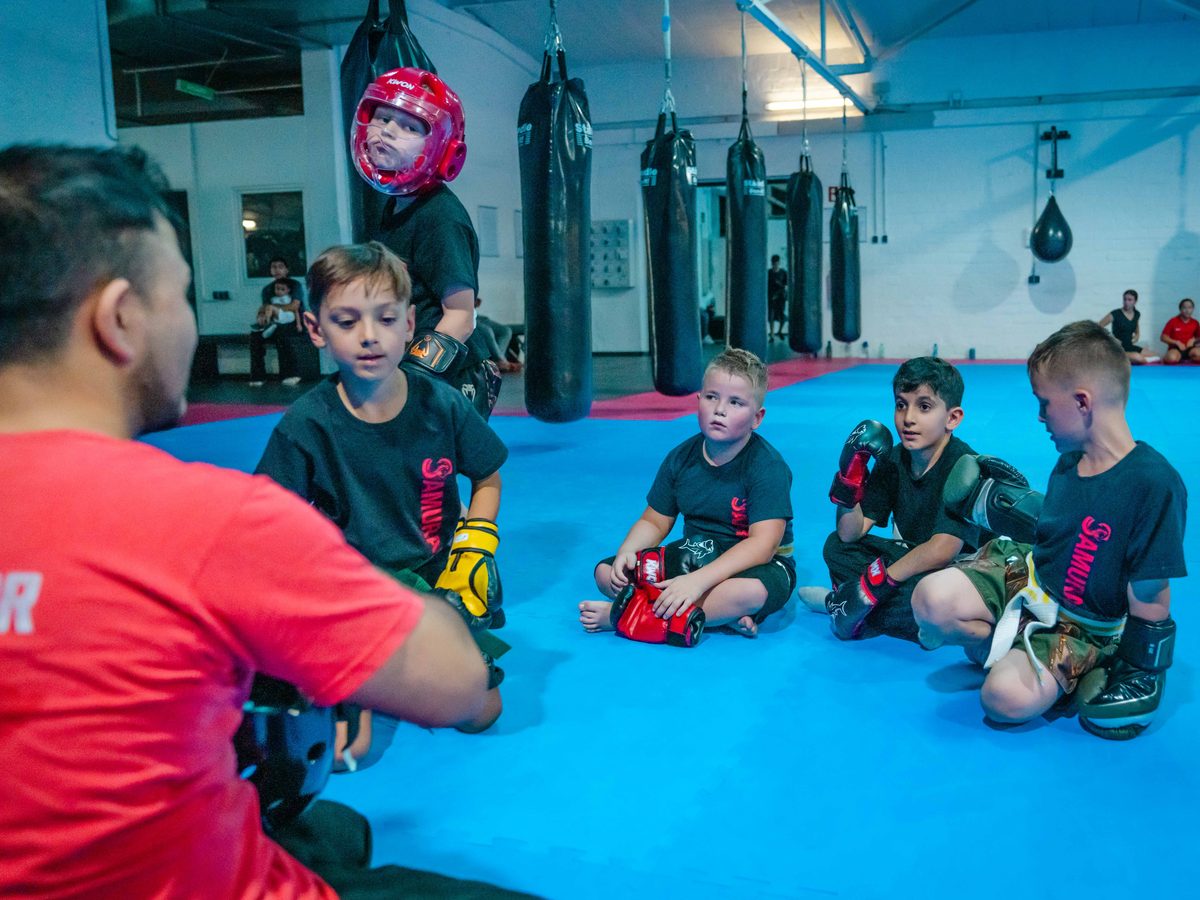 Kinder sitzen im Kreis auf der Matte, während der Trainer im roten Shirt mit ihnen spricht.