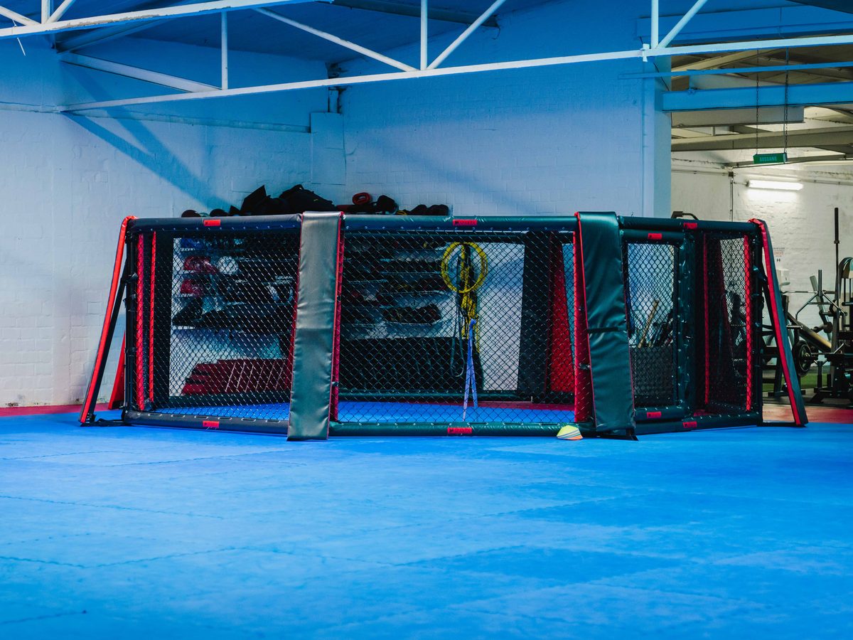 Der achteckige MMA-Käfig in der Trainingshalle, umgeben von blauer Mattenfläche und Sandsäcken.