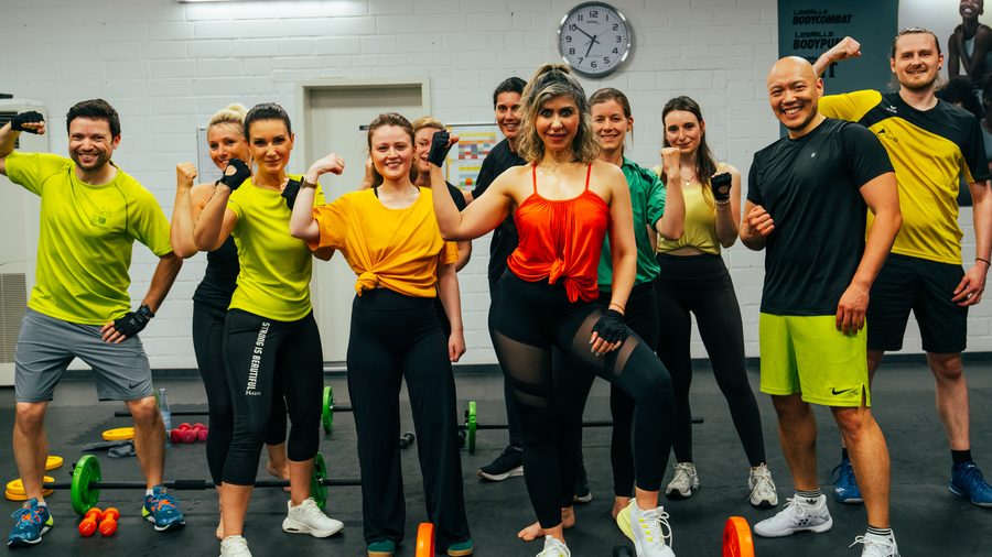 Gruppe von Body-Pump-Teilnehmerinnen und -Teilnehmern posiert mit Hanteln und Langhantelscheiben im Kursraum.
