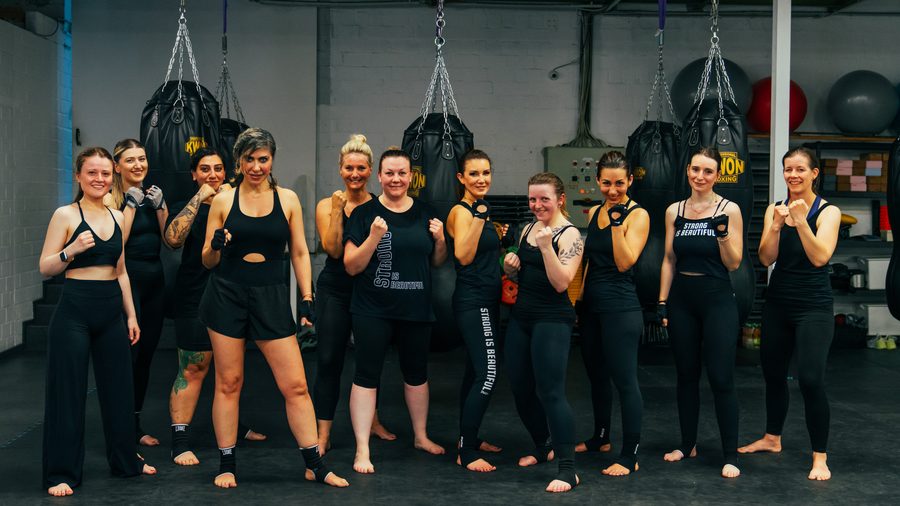 Elf Teilnehmerinnen des Frauen-Kickboxkurses stehen mit erhobenen Fäusten vor den hängenden Boxsäcken.