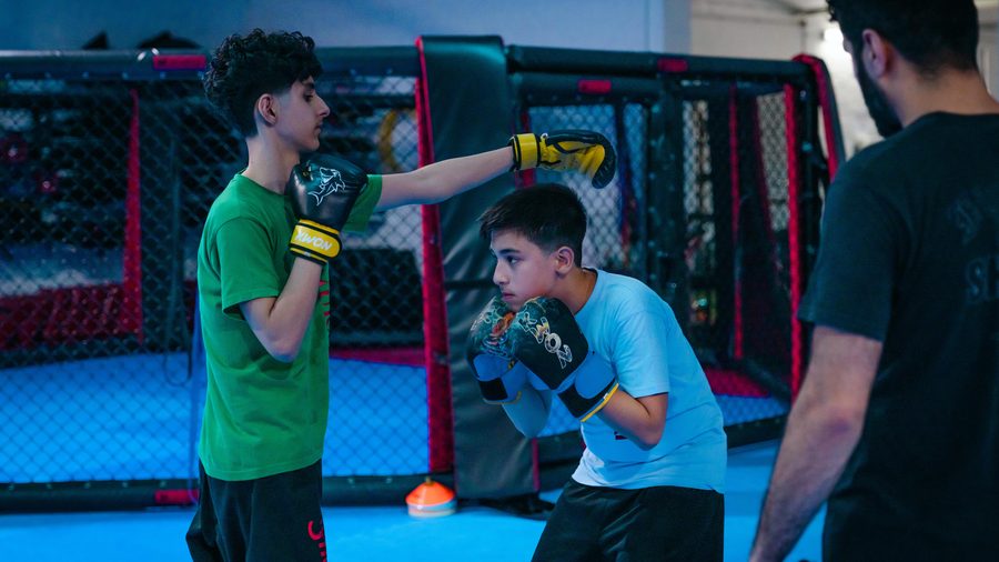 Zwei Jugendliche trainieren mit Boxhandschuhen und Pratzen im Bereich vor dem MMA-Käfig.