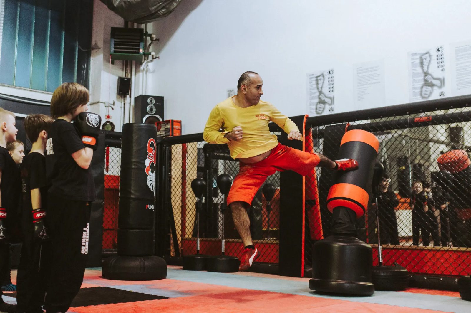 Probetraining 1 Der Sifu führt am Käfig einen hohen Kick gegen den Standsack aus, Kinder verfolgen die Übung.