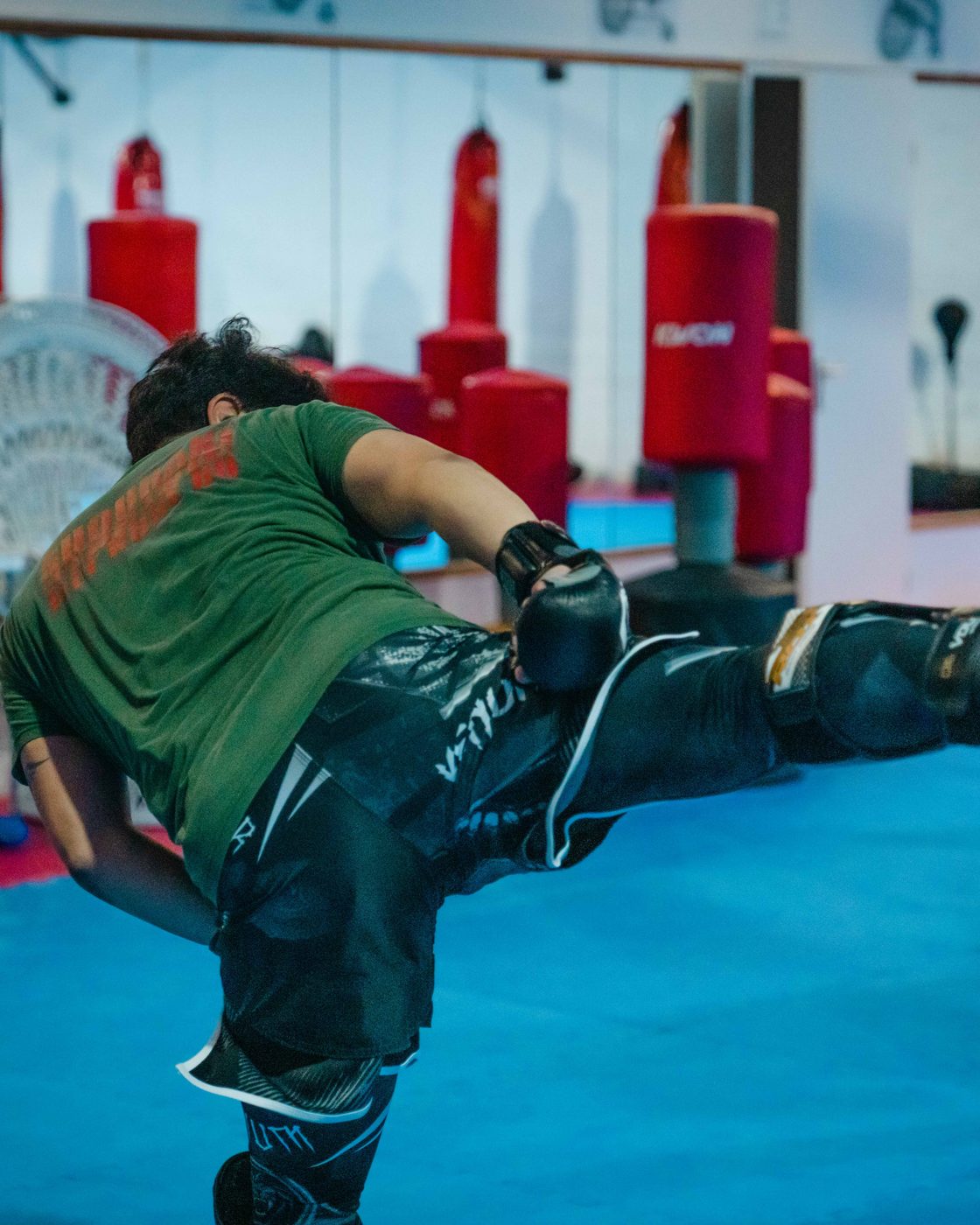 Trainierender führt einen hohen Seitkick gegen das Kickpad seines Partners aus, blau ausgeleuchtete Trainingshalle.