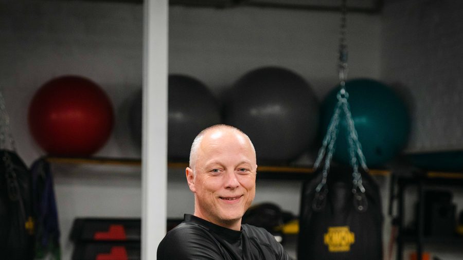 Trainer in schwarzer Trainingsjacke blickt vor Medizinbällen und Sandsäcken lächelnd in die Kamera.