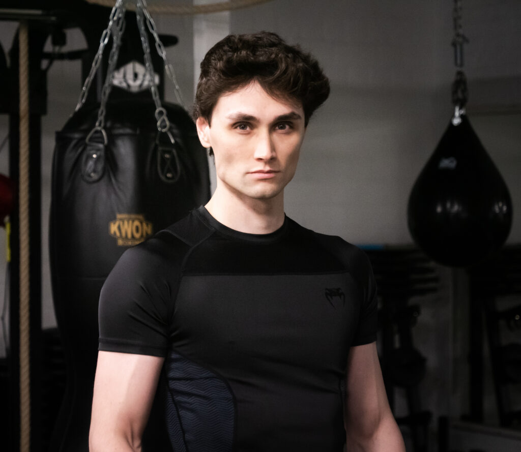 Junger Trainer im schwarzen Funktionsshirt vor hängenden Boxsäcken.