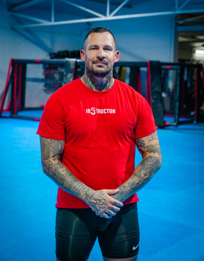 Trainer im roten Instructor-Shirt, Hände verschränkt, auf der blauen Matte.