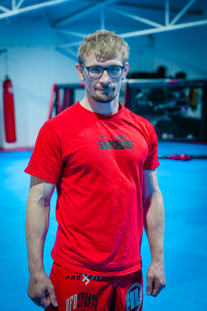 Trainer mit Brille im roten Shirt auf der blauen Matte vor dem Käfig.