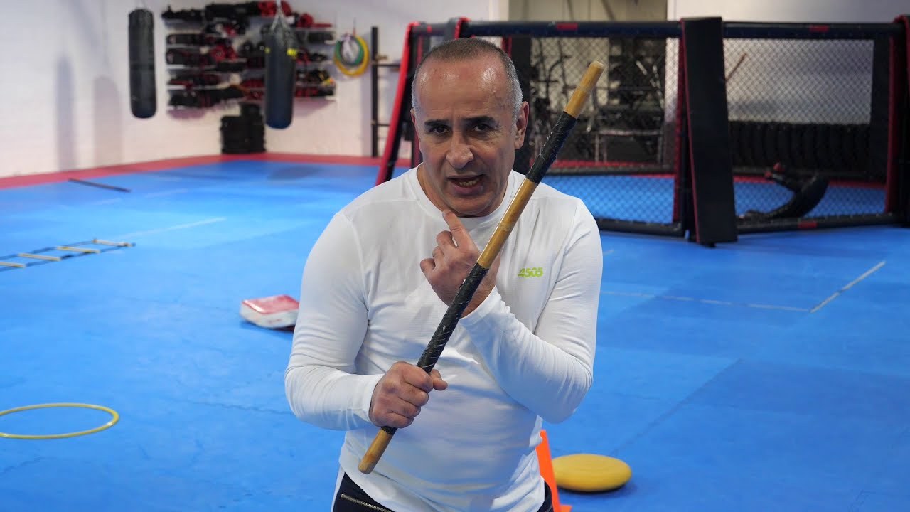 Still from the teaching video: the trainer holds a wooden stick with both hands in front of his chest and speaks to the camera.