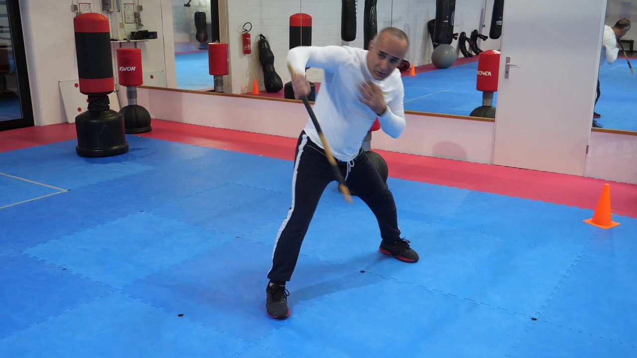 Still from the teaching video: the trainer swings the stick back, with standing bags and a mirror in the background.