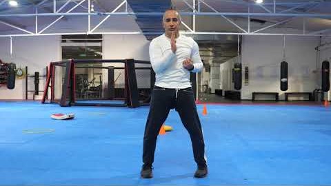 Still from the teaching video: the trainer stands on the blue mat in the training hall with his hands raised in front of his chest.
